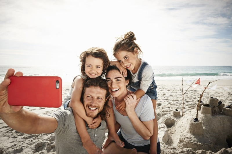 Lykkelig familie på fire tar en selfie på stranden, smilende mens de sitter tett sammen med et sandslott i bakgrunnen.