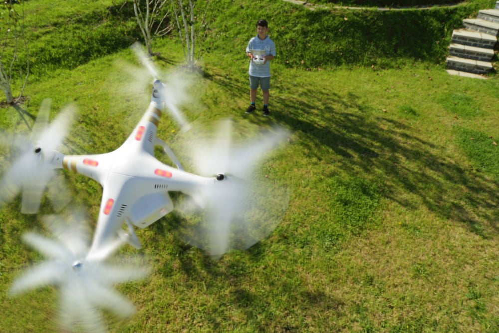 En ung gutt styrer en flyvende hvit drone på en solrik dag i en frodig hage. Dronen er i fokus med bevegelsesuskarphet på propellene, og viser teknologien i bruk i et naturlig og morsomt miljø.
