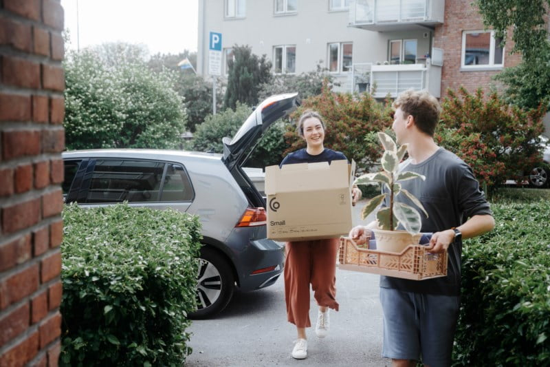 Et ungt par bærer flyttelass ut av en bil. Kvinnen smiler mens hun holder en pappeske, og mannen bærer en potteplante. De ser ut til å være midt i en flytteprosess i et boligområde.