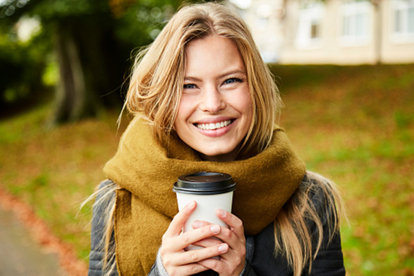 Smilende kvinne med stort skjerf holder engangskrus med kaffe